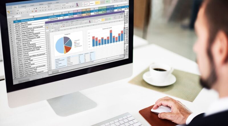 Person working at a desk on an iMac displaying Excel spreadsheets and charts (tables and bar/pie charts).