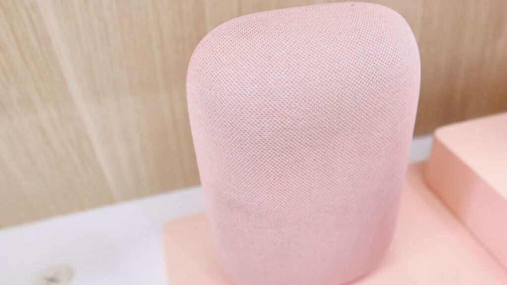 Pink cylindrical smart speaker with fabric mesh, close-up view on a pale pink surface.