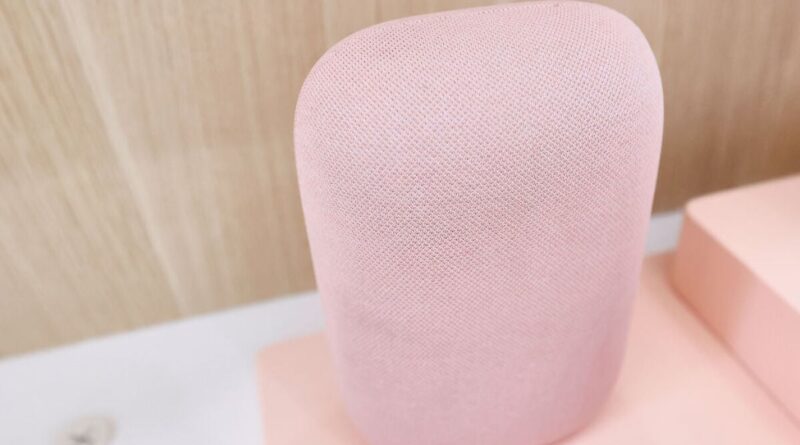 Pink cylindrical smart speaker with fabric mesh, close-up view on a pale pink surface.