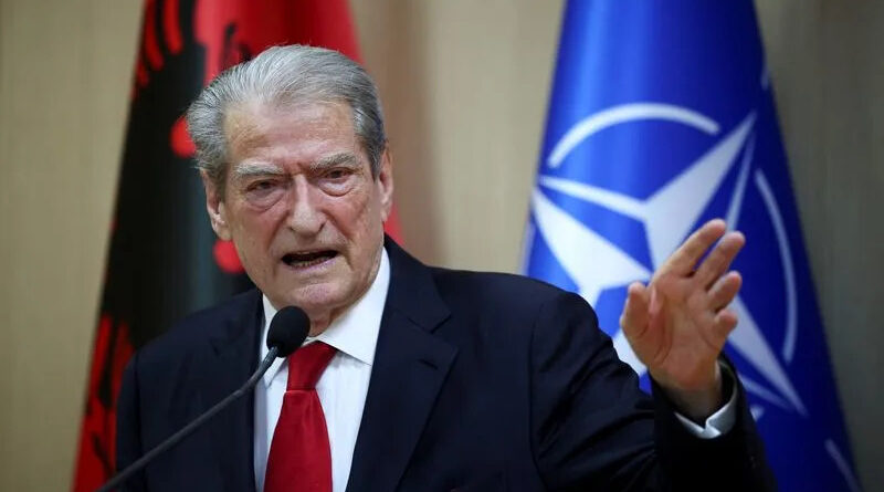 Older man in a dark suit and red tie speaks at a podium, with Albanian and NATO flags in the background