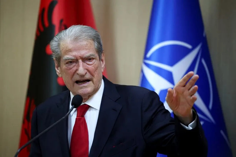 Older man in a dark suit and red tie speaks at a podium, with Albanian and NATO flags in the background