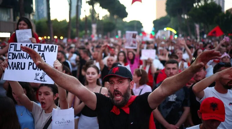 Crowd of protesters on a city street; man in a black shirt with a red scarf and cap raises arms at center.