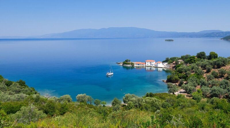 Tranquil coastal village on a turquoise bay with white houses and red roofs, a lone sailboat near the shore, and green hills in the foreground.