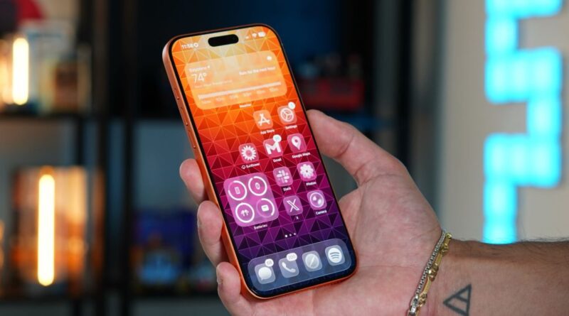 Close-up of a hand holding an iPhone with an orange case, showing a colorful app-filled home screen.