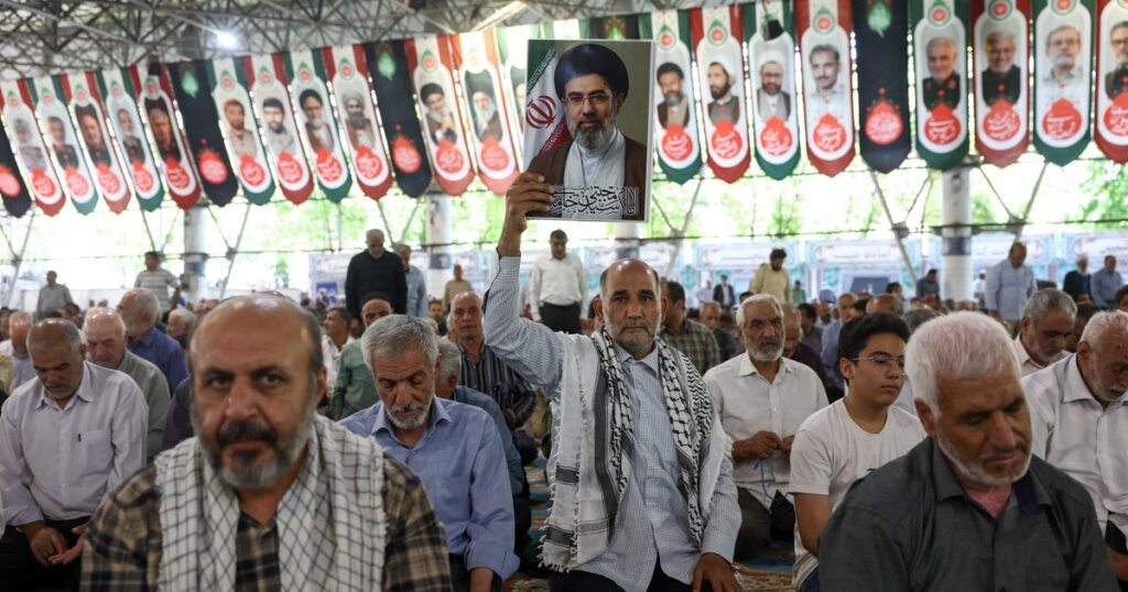 Man in a crowded hall raises a framed portrait of a religious leader during a bannered event.