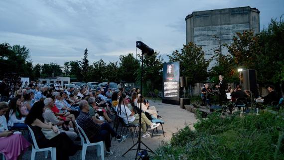 Outdoor evening concert: audience seated on plastic chairs facing a lit stage with musicians and a tall screen displaying a portrait or poster.