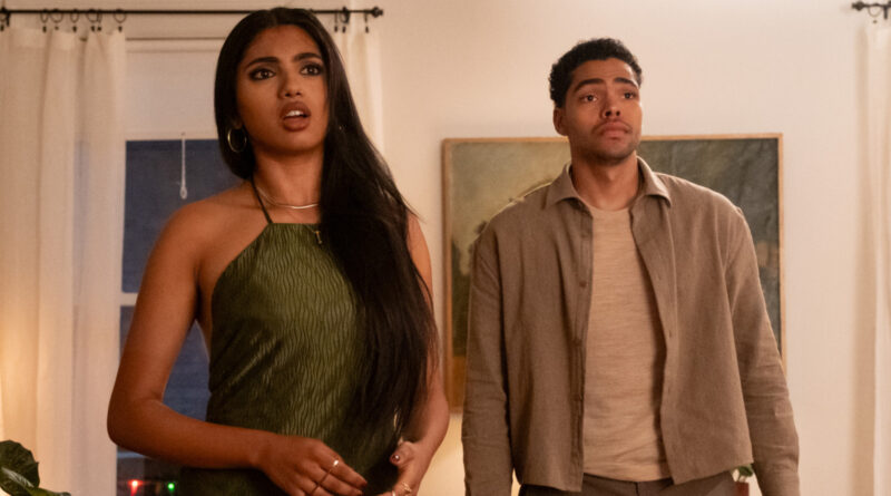 Two people stand indoors, the woman in a green halter dress looks surprised while the man in a beige jacket watches off-camera.