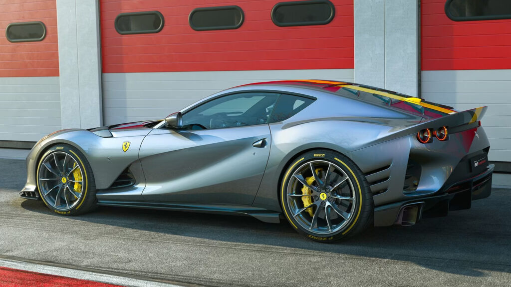 Side view of a metallic gray Ferrari sports car with yellow brake calipers parked in front of a red garage building.