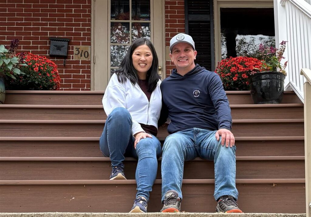 Smiling couple sits on brown wooden front steps of a brick house with red flower pots on either side. The woman wears a white zip-up jacket and jeans; the man wears a navy hoodie and jeans, with a light cap on.