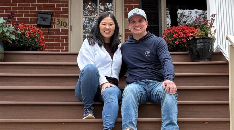 Smiling couple sits on brown wooden front steps of a brick house with red flower pots on either side. The woman wears a white zip-up jacket and jeans; the man wears a navy hoodie and jeans, with a light cap on.