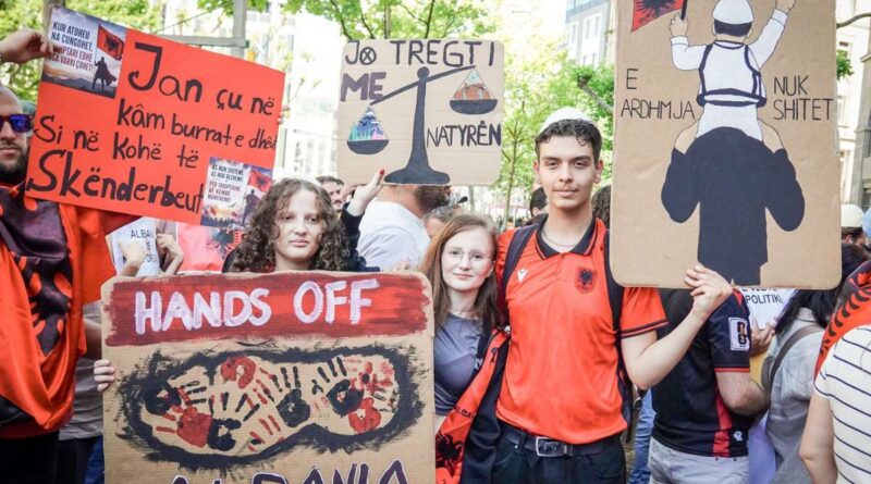Crowd of young protesters in orange shirts holding cardboard signs, including a large 'HANDS OFF' sign with red handprints, and other signs with symbols like scales of justice and a person riding on someone's shoulders.