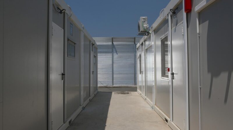 Narrow outdoor corridor between white industrial containers with doors and a wall-mounted AC unit, bright blue sky above.