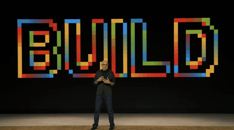 Man in a black shirt and jeans speaks on a stage in front of a large multicolored 'BUILD' backdrop on a black background.
