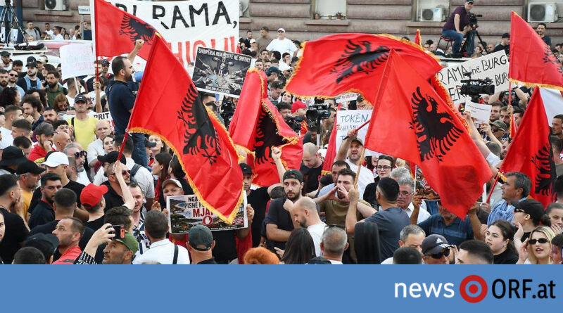 Dense crowd of protesters holding red Albanian flags with a black double-headed eagle; banners visible in the background; blue news banner at bottom.