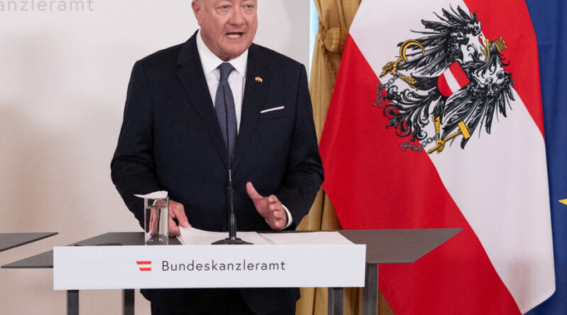 Bald man in a dark suit speaking at a podium with an Austrian flag behind him, Bundeskanzleramt backdrop behind.