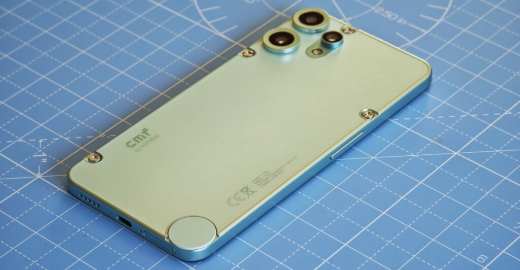 Back of a green smartphone with triple camera module in top-left corner on a blue grid cutting mat, with small screws visible and a circular sensor at the bottom edge.