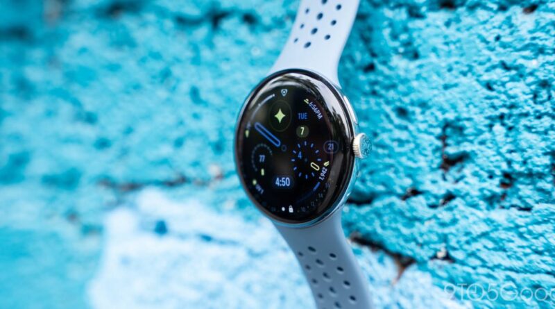 Round smartwatch on a light gray perforated strap against a blue textured wall, showing a colorful digital watch face with apps and time.