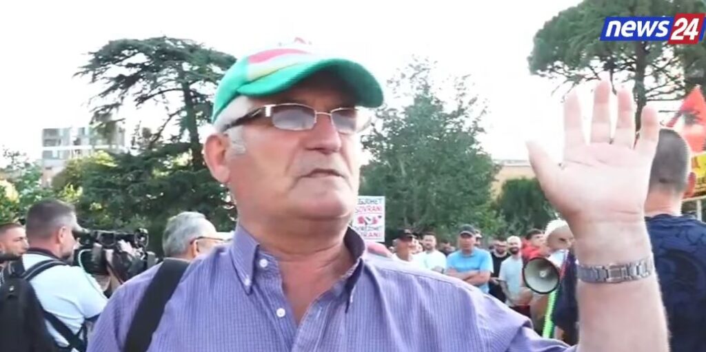 Older man in a purple shirt and green cap raising his hand at a protest, with a crowd and cameras in the background; news24 logo top-right.
