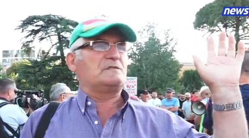 Older man in a purple shirt and green cap raising his hand at a protest, with a crowd and cameras in the background; news24 logo top-right.