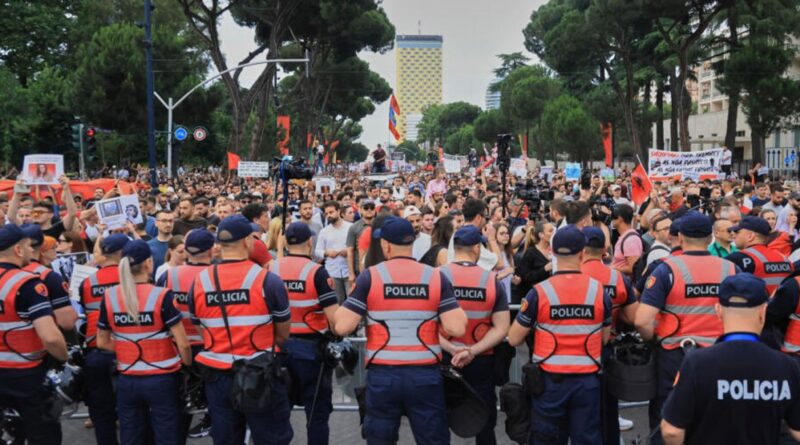 Line of police in orange vests facing a large protest crowd with signs and flags in a city plaza. They form a barrier between demonstrators and the street.
