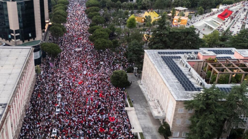 Aerial view of a massive protest march down a city street, thousands wearing red and white, flanked by buildings and trees in a park area to the side.