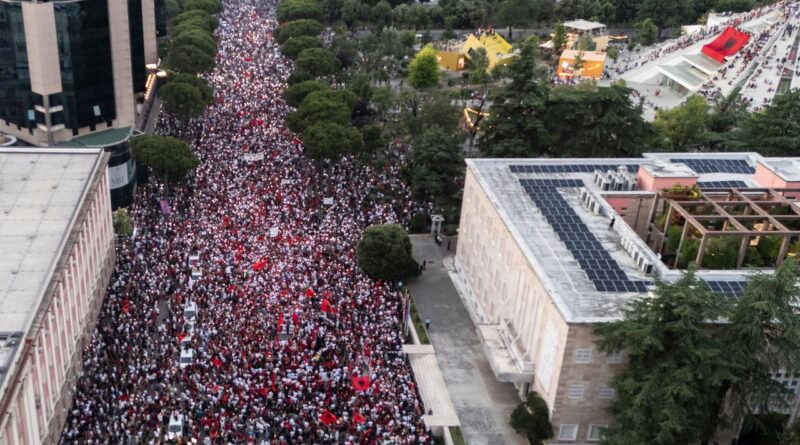 Aerial view of a massive protest march down a city street, thousands wearing red and white, flanked by buildings and trees in a park area to the side.
