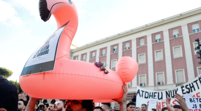 Bright pink inflatable flamingo held above a protesting crowd, with a banner strapped to it and a large banner with text in the background.