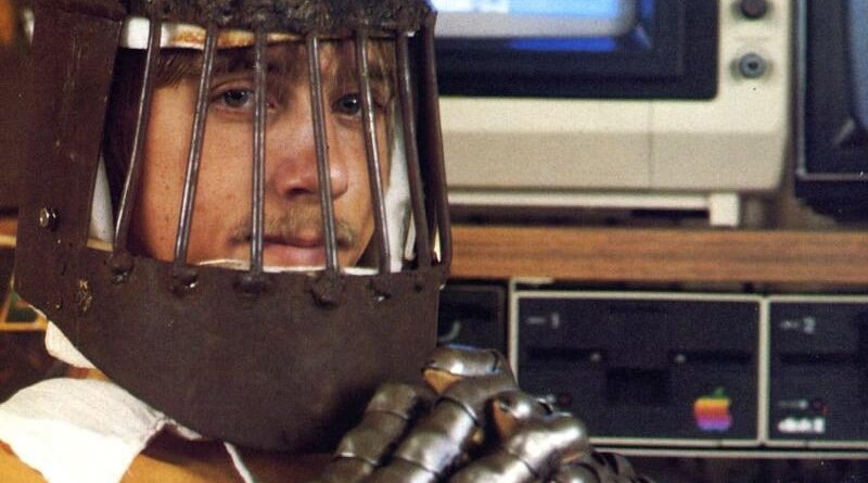 Person wearing a metal cage-like helmet with face bars sits in front of vintage computer equipment.