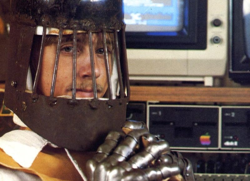 Person wearing a metal cage-like helmet with face bars sits in front of vintage computer equipment.