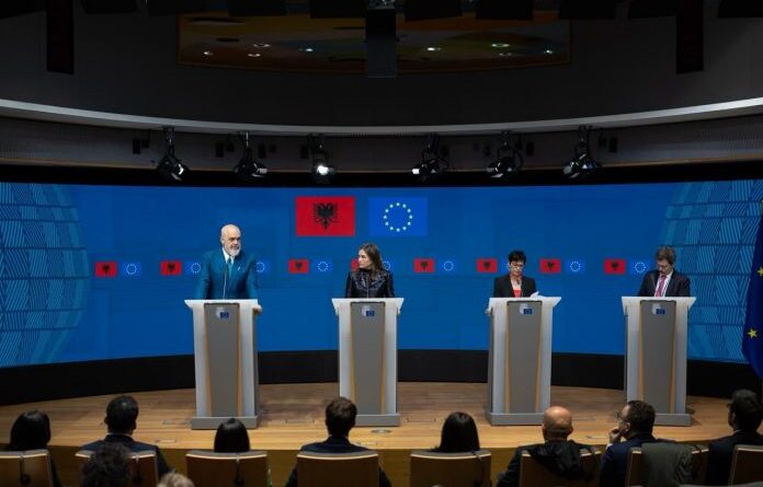 Four speakers stand at podiums on a stage during a press conference, with Albanian and European Union flags on a blue backdrop and an audience in front.