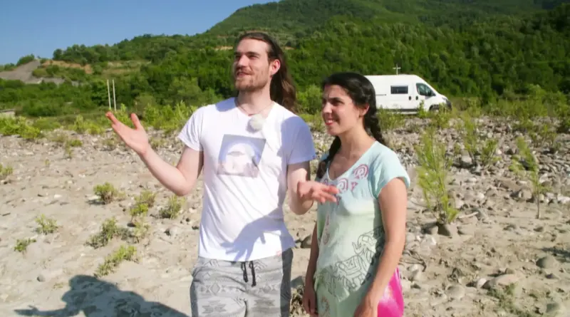 Two people standing on a rocky outdoor area with a white van and green hillside in the background, talking or explaining something.