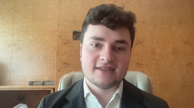 Young man in a dark blazer and white shirt sits in a chair, looking at the camera during a video call.