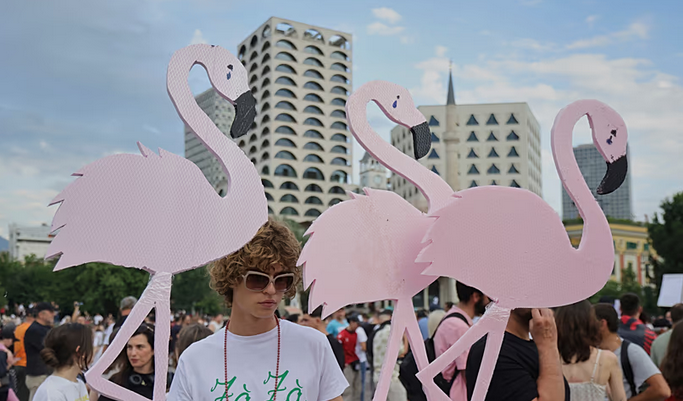 Person wearing sunglasses stands among oversized pink cardboard flamingos at a crowded city street event with modern buildings in the background.