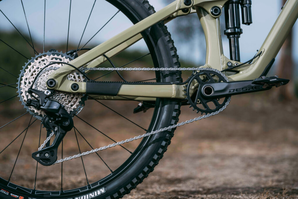 Close-up of a mountain bike rear drivetrain: cassette, chain, derailleur, and beige frame.