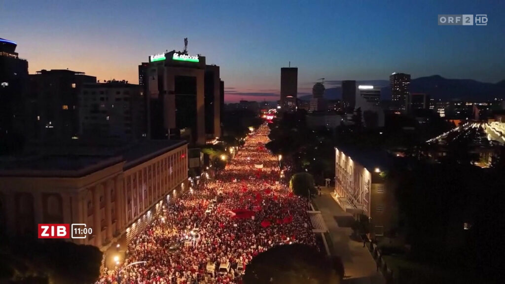 Aerial view of a crowded city street at dusk with thousands of protesters marching along a well-lit avenue between buildings.",