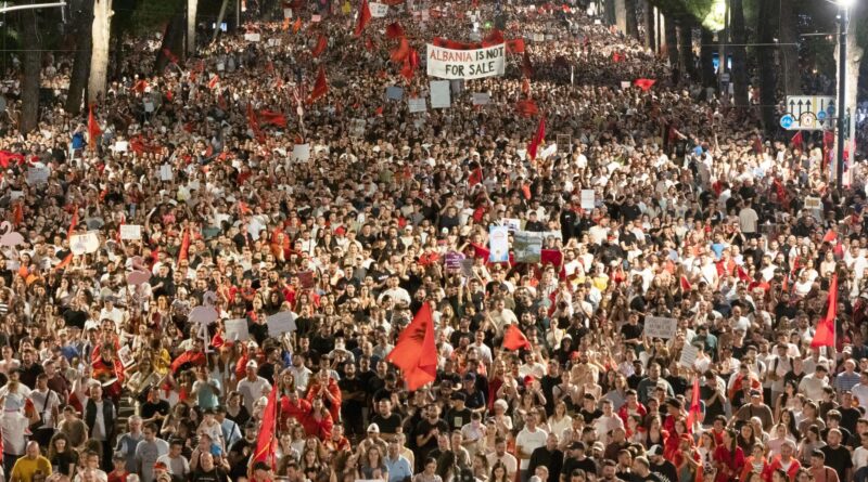 Huge protest crowd filling a wide street with red flags and signs; a banner reads 'Albania is not for sale'.