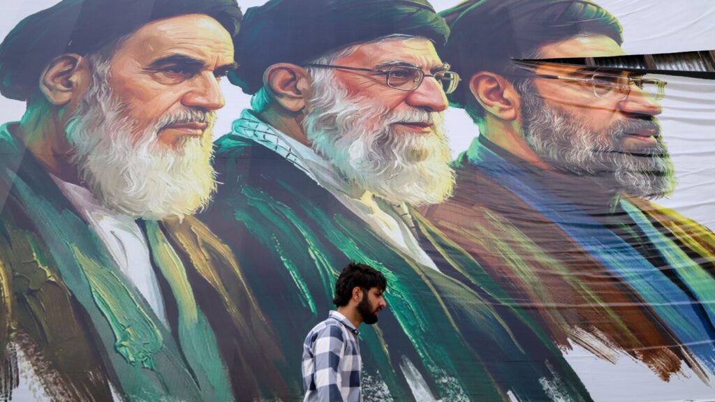 Man with a beard walks past a large colorful mural of three bearded men in religious attire.
