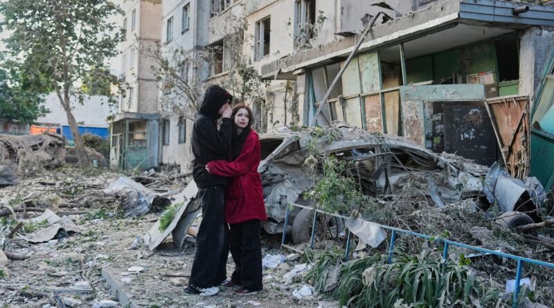 Couple in dark clothing embrace amid rubble and a ruined building in a war-torn street