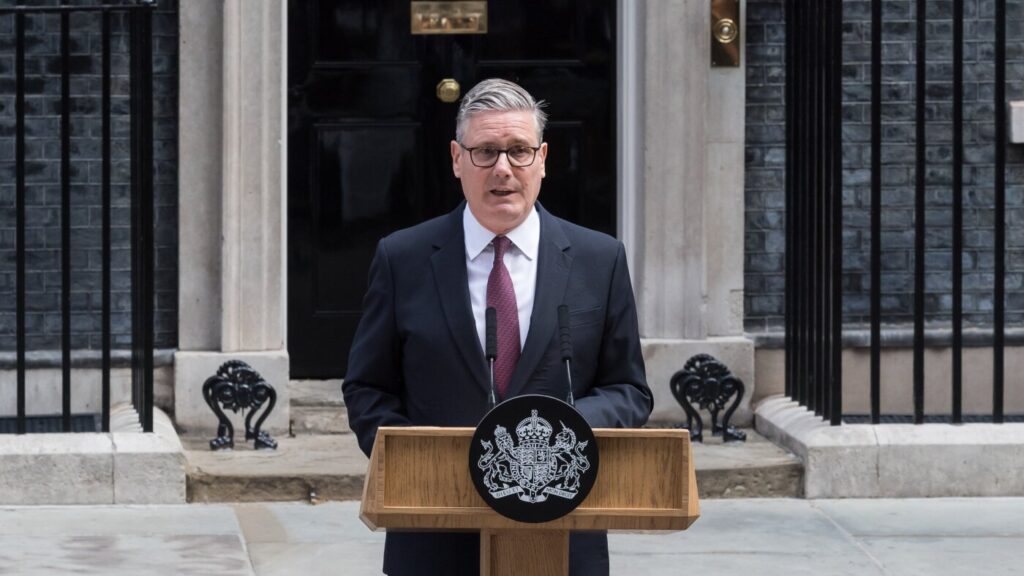 Man in a suit speaks at a wooden podium with a UK crest emblem, outside a government building.