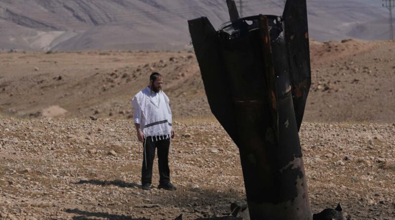 Bearded man in a white prayer shawl stands beside a large burnt aircraft fuselage wreck in a desert landscape