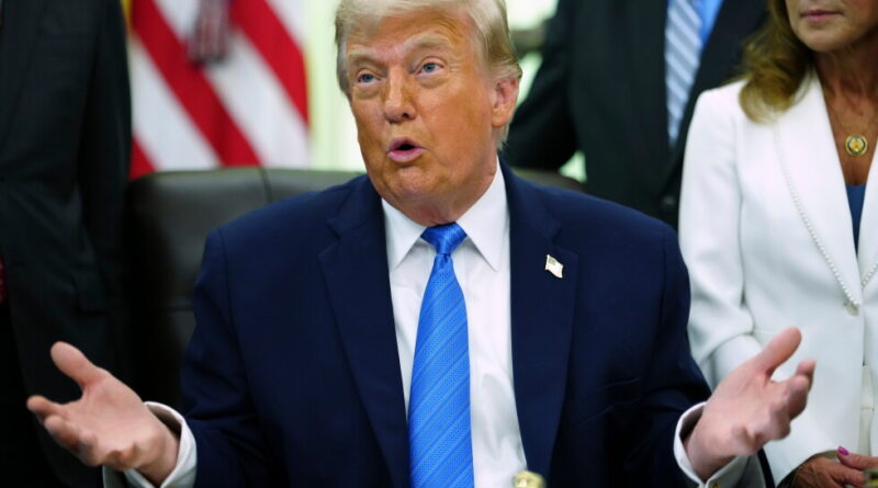 Man in a navy suit and bright blue tie speaks with open hands at a conference table, American flag in the background.