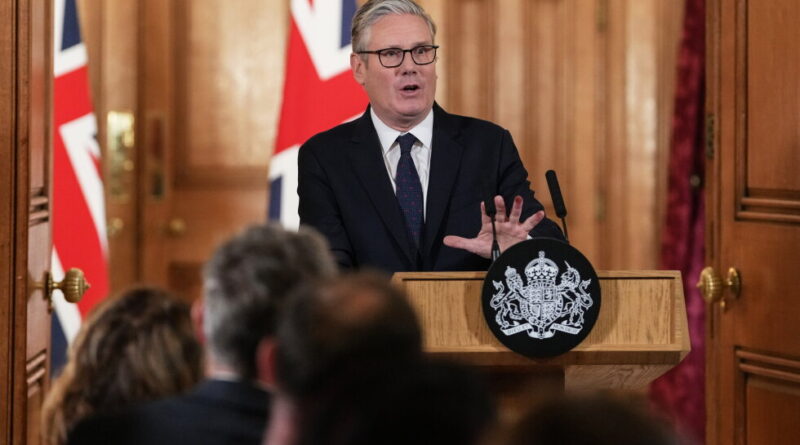 A man in a dark suit and glasses speaks at a wooden podium with a crest, flags behind him, addressing an audience at a formal briefing.