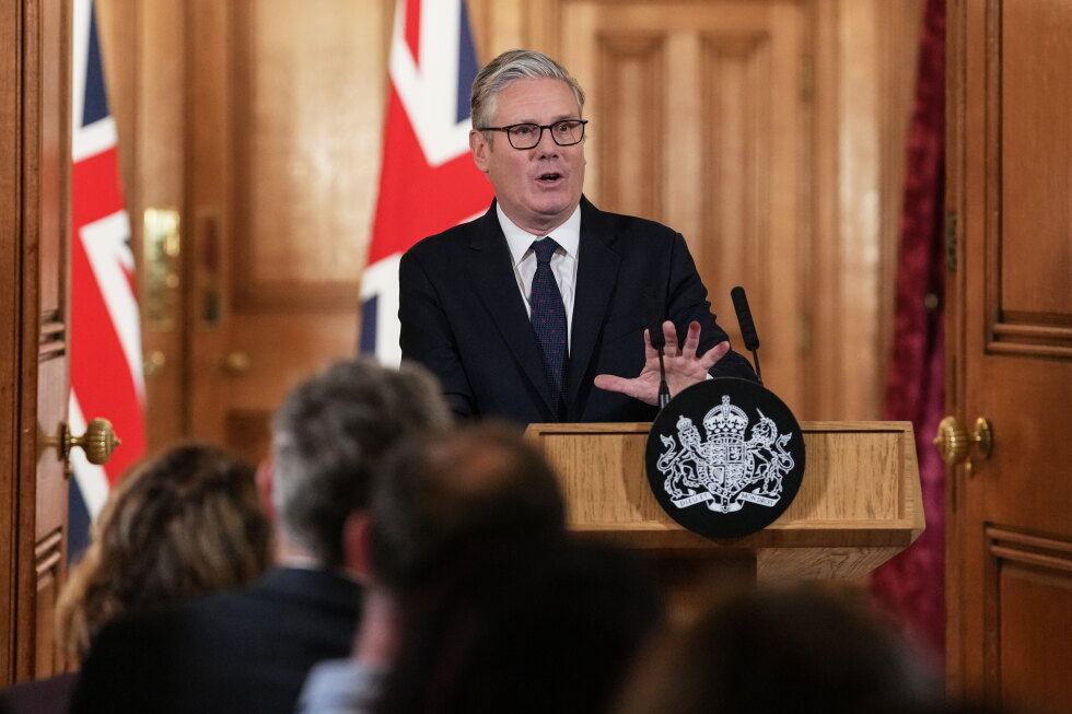 A man in a dark suit and glasses speaks at a wooden podium with a crest, flags behind him, addressing an audience at a formal briefing.