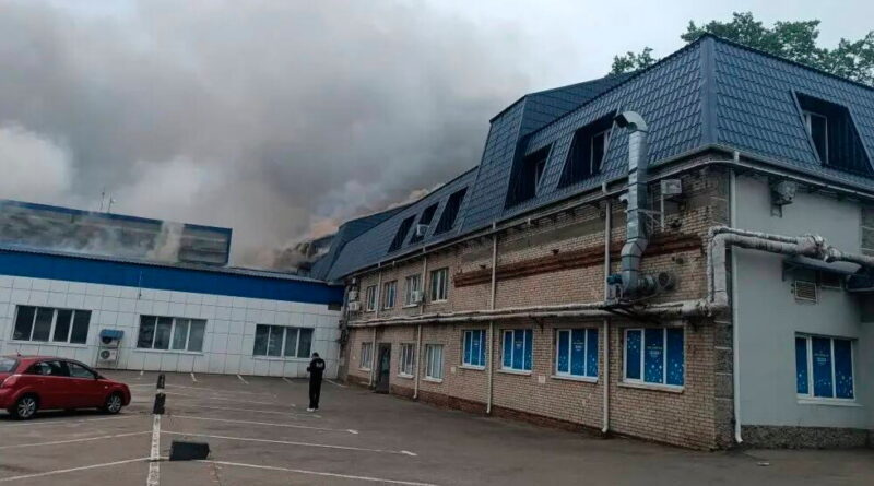 Brick building with a blue metal roof and heavy smoke rising from the left side; a person stands in a parking lot nearby.