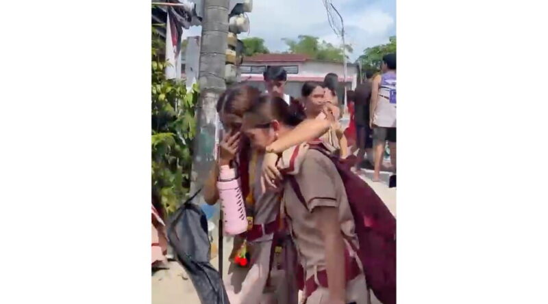 Two students in uniform comfort a crying peer on a sunny street, one arm around the other near a utility pole and backpacked exterior scene.