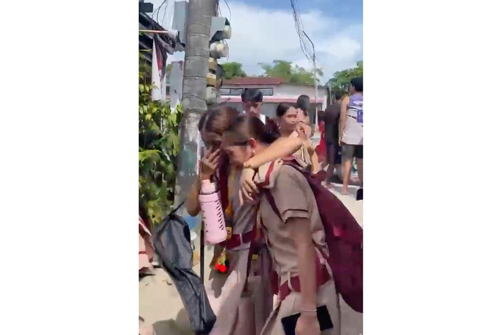 Two students in uniform comfort a crying peer on a sunny street, one arm around the other near a utility pole and backpacked exterior scene.