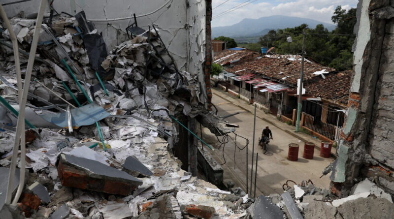 Rubble and collapsed walls of a building overlook a street with a man riding a motorcycle beyond the debris.