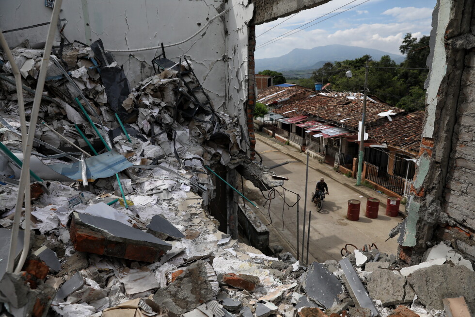 Rubble and collapsed walls of a building overlook a street with a man riding a motorcycle beyond the debris.