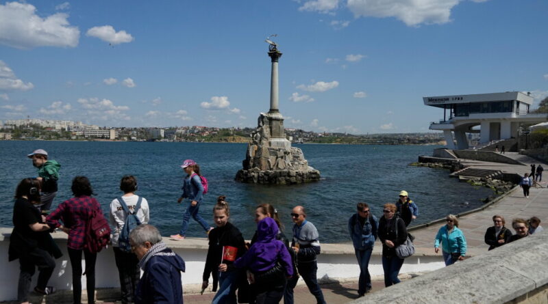 Crowded seaside promenade with a tall monument on a rocky pedestal in the water and people strolling along the waterfront.