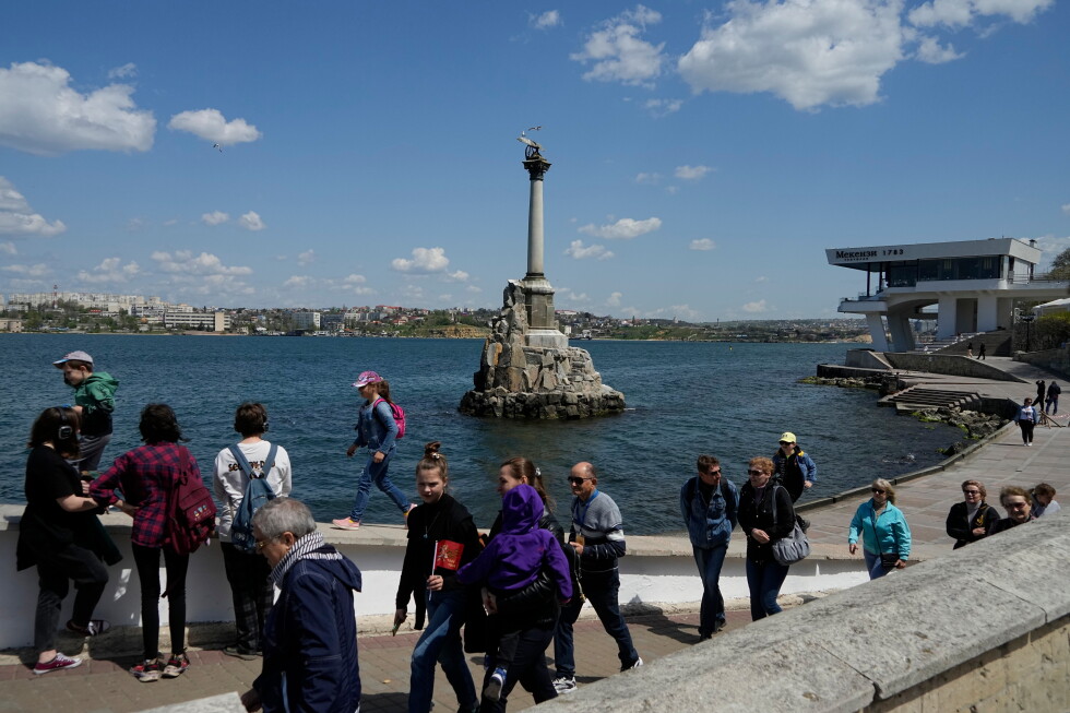 Crowded seaside promenade with a tall monument on a rocky pedestal in the water and people strolling along the waterfront.